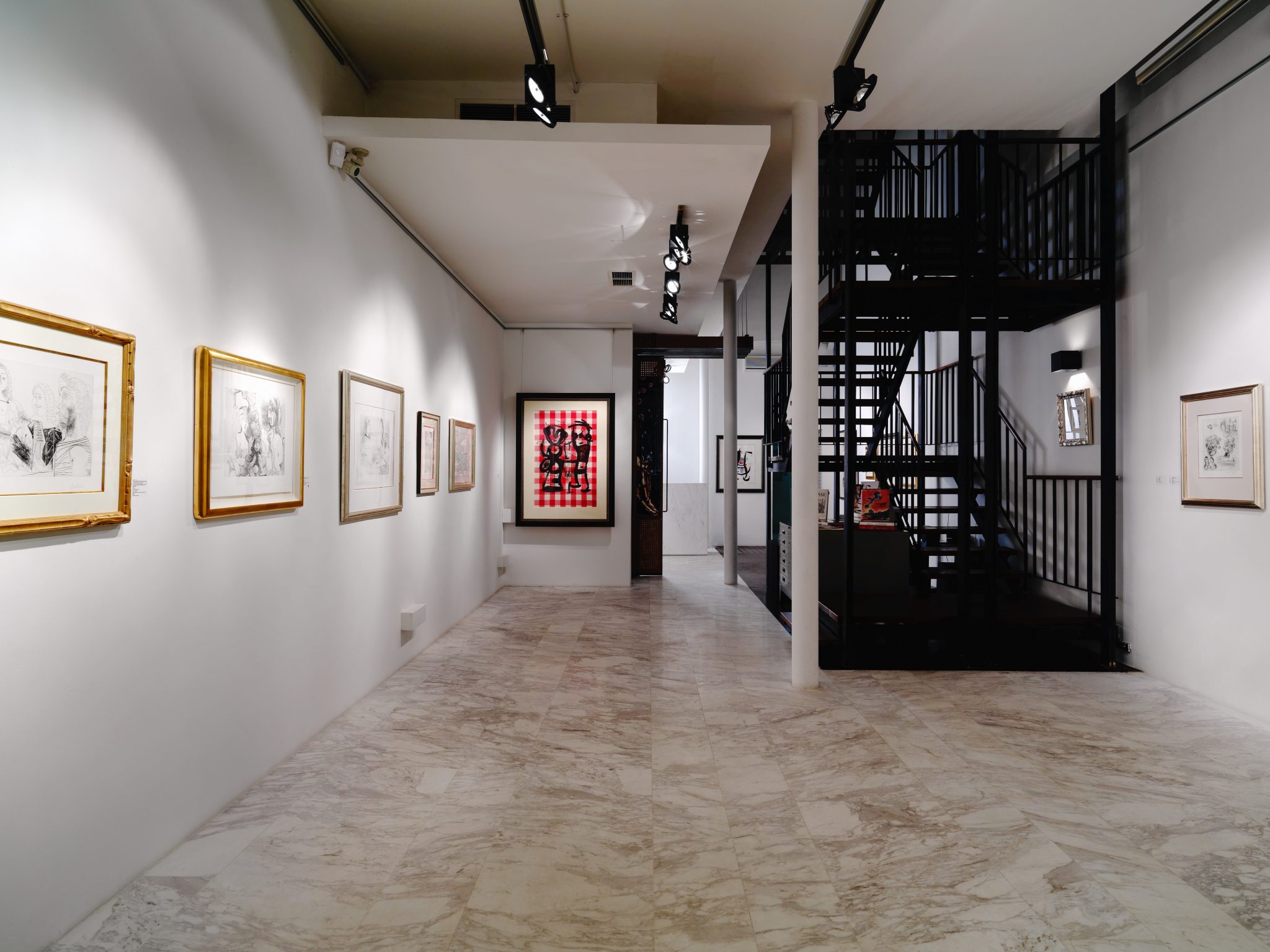 FOST Gallery by PIU: white gallery wall of framed works beside a black steel staircase on marble flooring